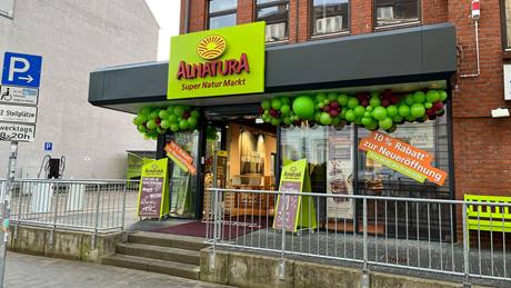 Der Eingang des Alnatura Super Natur Markts in Kiel in der Kirchhofallee am Tag der Neueröffnung mit Luftballons und Plakaten geschmückt. Rechts am Eingang steht eine grüne Sitzbank.