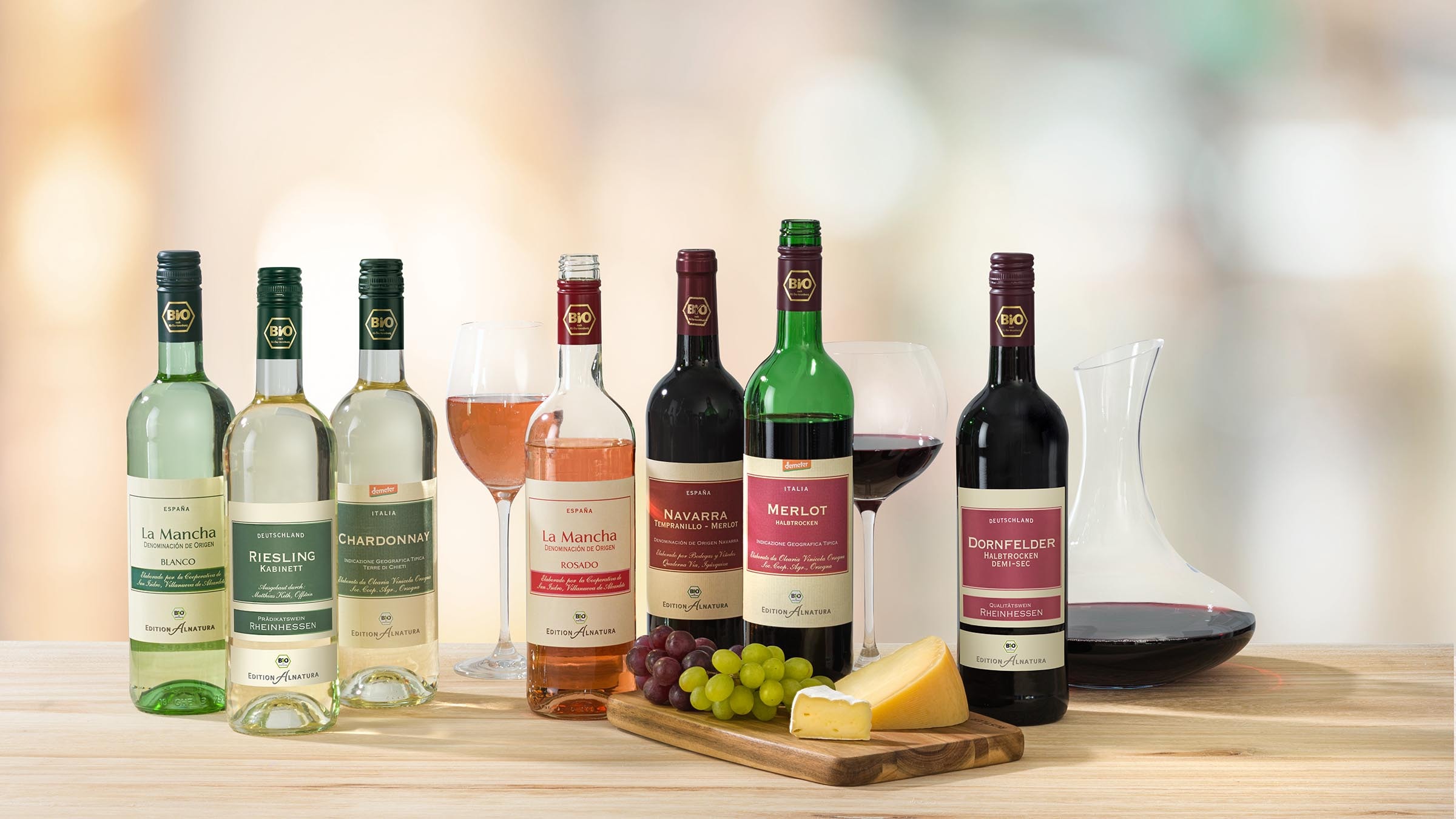 Auf einem Holztische stehen insgesamt sieben Flaschen Wein. Links drei Weinflaschen mit unterschiedlichen Weißwein-Sorten, die teils geöffnet sind.Rechts daneben ein Glas und eine geöffnete Flasche mit Roséwein. In der Mitte des Bildes stehen zwei Rotwein-Flaschen, eine davon ist geöffnet und ein Glas mit Rotwein ist zu sehen. Am rechten Bildrand steht eine weitere geschlossene Rotwein-Flasche und eine Glaskaraffe (Dekantierer) mit eingegossenem Rotwein. Im Vordergrund ist ein Holzbrett mit Weintrauben und einem Stück Käse. 