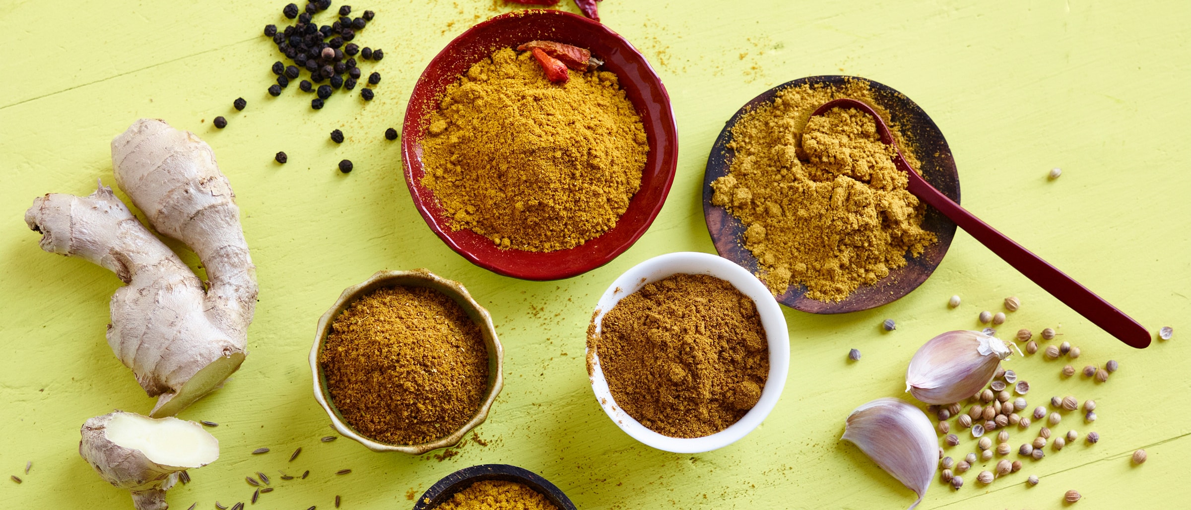 verschiedene Currypulver Sorten in Schälchen, Knoblauchzehen, schwarze Pfefferkörner, bunte Pfefferkörner