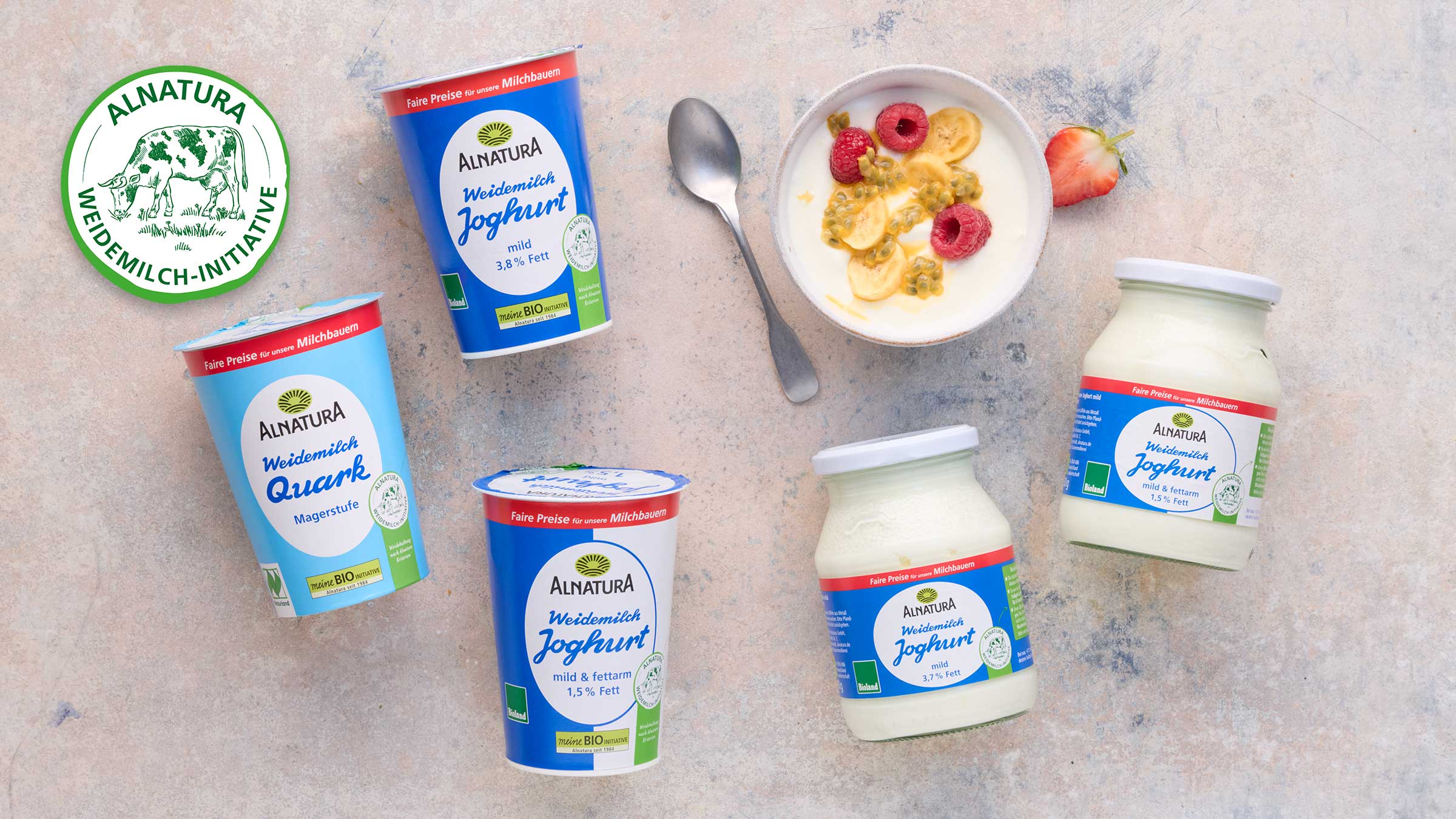 Das Bild zeigt verschiedene Produkte der Marke Alnatura: Joghurt (mild, 3,8% Fett; mild & fettarm, 1,5% Fett) und Magerquark.  Die Produkte sind auf einem hellen Untergrund arrangiert, neben einer Schale Joghurt mit Früchten (Himbeeren, Banane, Passionsfrucht) und einem Löffel.  Im Hintergrund ist ein Logo der "Alnatura Weidemilch-Initiative" zu sehen, welches auf faire Preise für Milchbauern hinweist.  Das Bild wirkt werblich und soll die Produkte und die Initiative präsentieren.